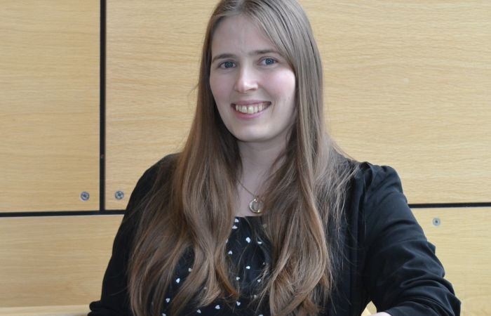 Photo of a women standing in a well lit stair well with her elbow leaning on the banister. The women is wearing a black suit top and is smiling.