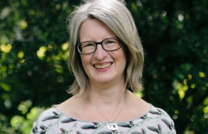 Photo of a woman standing in front of dark green leaves, wearing a green and cream top and glasses,smiling